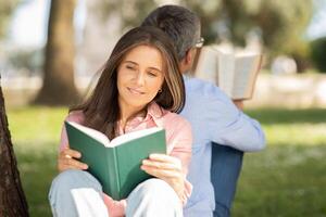 A woman enjoys a book while seated against a tree, her back to a man who is also reading. The warm sunlight filters through the branches, creating a peaceful atmosphere in the park. photo