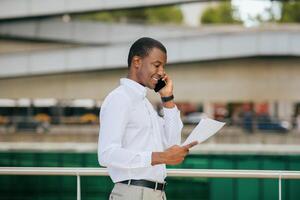 Black man wearing a white long-sleeved shirt is talking on his phone while reviewing documents. He is standing outside in front of a modern building with a green railing. photo