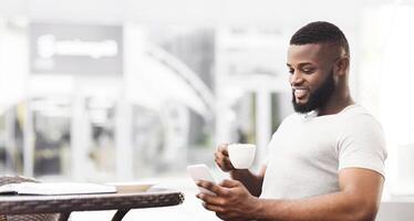 A man with a beard sits at a cafe table, smiling while looking at his smartphone and holding a cup of coffee. photo