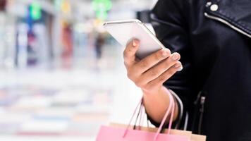 A woman standing while holding a pink shopping bag in one hand and a cell phone in the other. She appears to be engaging in a shopping activity while using her phone, cropped photo