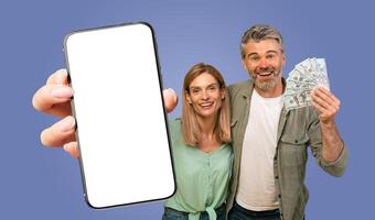 A couple is shown smiling and holding a smartphone with a blank screen and cash in front of a blue background. The woman is holding the smartphone while the man is holding a fan of cash. photo