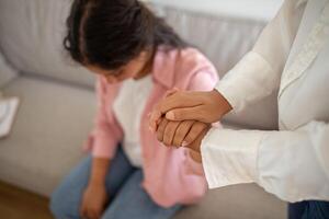 A woman in a white shirt holds the hand of another woman sitting on a sofa, offering comfort and support. The woman receiving comfort has her head down, suggesting sadness or distress. photo