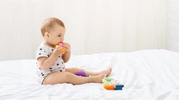 A baby girl wearing a white onesie sits on a white bed with a white comforter. The baby is playing with colorful plastic toys. The babys face is turned to the side and she is looking at the toy photo