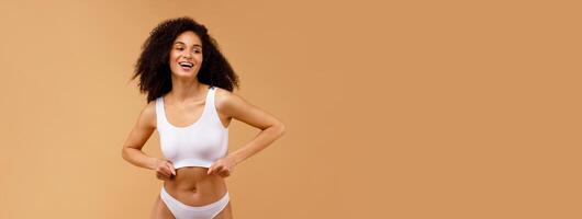 Slim african american woman in white underwear top bra and panties standing isolated over beige studio background, posing and smiling, free space photo
