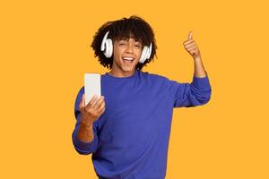 This image shows a man with a joyful expression while wearing headphones and listening to music on his phone. He is holding his phone in his left hand and giving a thumbs up with his right hand photo