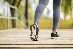 A person is taking a morning jog on a wooden pathway through a park. Wearing athletic gear, the runner steps confidently forward in black running shoes against a backdrop of green trees. photo