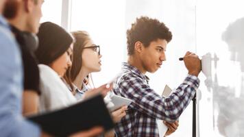A diverse group of students standing in front of a whiteboard, engaged in writing and discussing ideas. The whiteboard is filled with diagrams, bullet points, and equations photo