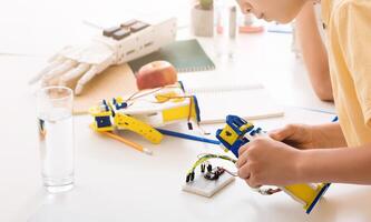 A young student sits at a desk, focusing intently on building a robotic hand. The hand is a bright yellow and blue, and the student is holding it with both hands, studying the wires and circuitry photo