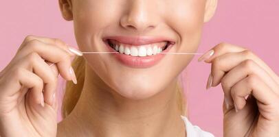 A close-up of a woman flossing her teeth with white floss. Her mouth is open and she is smiling, showing her white teeth. The background is a solid pink color. photo