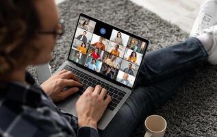 A man sits on the floor with his legs crossed, using a laptop to participate in a group call. The laptop screen shows a grid of multiple participants in the call. photo
