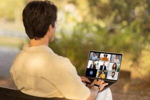 A man sits on a bench in a park, engaging in a conference on his laptop. The natural surroundings and daylight create a serene environment while he attends the virtual meeting photo