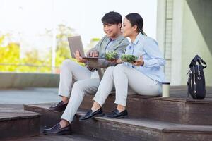 Seated on steps outside an office building, Chinese man and woman dressed in business casual attire are engaged in a collaborative task using a laptop, have lunch break photo