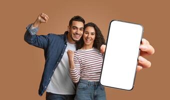 This image shows middle eastern couple standing in front of a brown background. They smiling and excited, with their fists raised in the air. The woman is holding a smartphone with a blank screen. photo