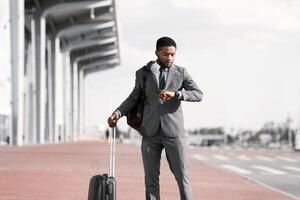 A professional black man with a suitcase is checking his watch outside an airport. The mood is efficient and on-the-go, with the modern airport setting creating a sense of urgency. photo