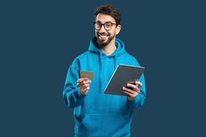 A cheerful man, dressed in a blue hoodie, holds a credit card in one hand and a tablet in the other. He appears excited about online shopping, showcasing the convenience of digital transactions photo