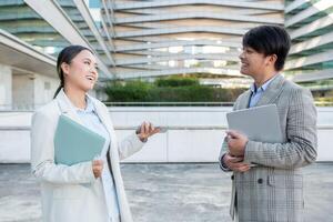 Asian woman and man are conversing engaged in front of a contemporary office structure. The woman is holding a tablet and appears to be sharing insights, while the man listens intently photo