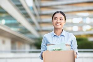 Excited Asian businesswoman is standing while holding a cardboard box. She appears focused and determined, possibly moving office or transporting items photo