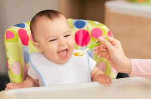 A joyful baby boy sits in a colorful high chair, eagerly opening their mouth as a caregiver feeds them with a spoon of baby food in a bright, cheerful kitchen. photo
