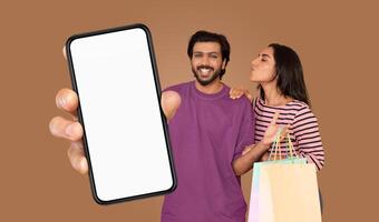 A man and a woman stand together in front of a brown background, holding a smartphone with a blank white screen and shopping bags. photo