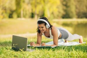 A woman in athletic wear is seen doing a plank exercise on a yoga mat while watching a laptop, with headphones on in a sunny park. photo