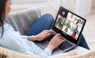 A woman is sitting on a couch, engaging in a virtual meeting with multiple participants displayed on her laptop screen. She appears focused and involved in the discussion photo