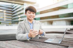 Asian man wearing a gray blazer sits at a wooden table outside, using his smartphone while a laptop is to the right. He is looking at his phone and smiling. The setting appears to be a city photo
