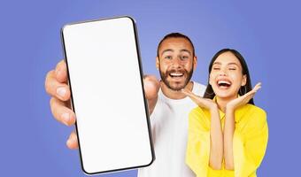 A man and a woman are standing in front of a blue background, both smiling and looking at the camera. The man is holding a smartphone with a blank screen in his hand, and the woman is smiling photo