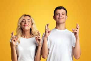 Make a wish. Smiling millennial couple crossing fingers for luc standing with closed eyes over yellow background, panorama with copy space photo