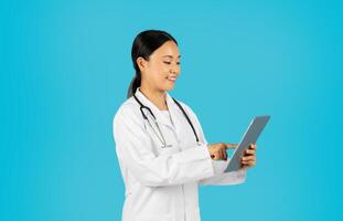 Asian woman doctor, wearing a white lab coat and a stethoscope, stands in front of a blue background, smiling while using a digital tablet. photo