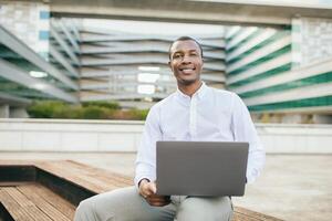 African American man sits on a wooden bench outside of a modern building, working on his laptop. He is smiling and looking directly at the camera. He appears to be focused on his work. photo