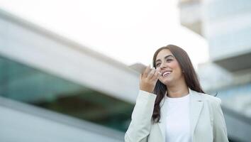A young woman in a white blazer smiles as she uses the voice recognition feature on her smartphone. She stands outside in an urban setting, with buildings in the background, copy space photo