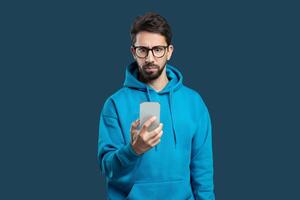 A young man wearing a blue hoodie is holding a smartphone in one hand while sporting a serious expression. He appears to be engaged in a focused activity, possibly examining content on the device photo