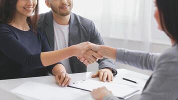 A couple sits at a table with a financial advisor, shaking hands in agreement. The couple is dressed casually while the advisor is dressed professionally, cropped photo