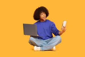 A young guy is seated on the floor, elegantly crossed-legged, engaged in a joyful interaction with a smartphone while using a laptop. The bright yellow background photo