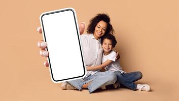 African American mother and her young son are sitting on the floor, smiling while holding a blank smartphone towards the viewer. They are dressed casually, creating a warm and playful atmosphere. photo