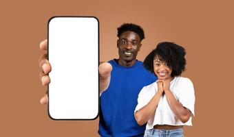 African American young man and woman stand in front of a brown background, the man holding a smartphone with a blank screen in front of him. Both are smiling directly at the camera photo
