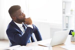 Thoughtful afro businessman making business solution at workplace, touching chin and looking aside, empty space photo