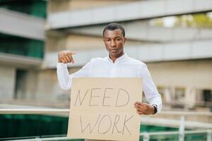 African American young man in a white shirt stands in front of a building, holding a cardboard sign that reads Need Work. He is looking directly at the camera with a serious expression. photo
