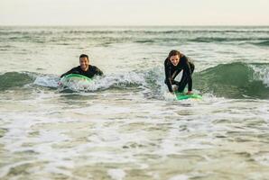 A couple in wetsuits are enjoying a day surfing on the ocean. The man is lying on his board, while the woman is kneeling on hers. They are both smiling and look like they are having a lot of fun. photo