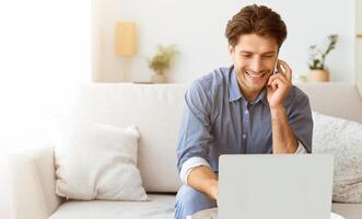 A man smiles while sitting on a couch, using a laptop and talking on the phone. He appears to be relaxed and happy. photo