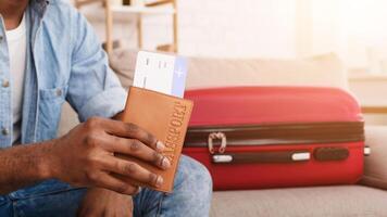 Cropped of black man is seated, displaying a passport and boarding pass, suggesting he is prepared for an upcoming journey. A red suitcase next to him photo