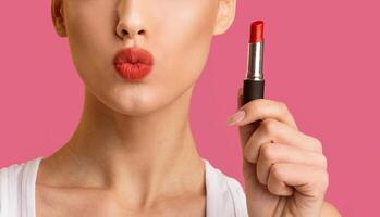 Cropped of a woman holding a red lipstick in her right hand. The woman is wearing a white tank top and her lips are painted red. The background is a solid pink color. photo