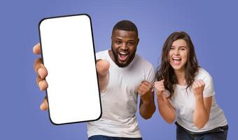 Multiracial couple standing in front of a purple background. The man is holding a smartphone with a blank screen towards the camera, while the woman is smiling and clenching fists photo