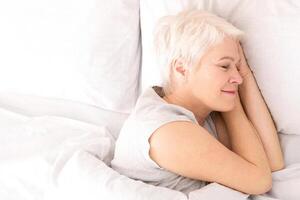 A woman is lying down in bed, covered by crisp white sheets. She appears relaxed and comfortable, with her eyes closed, possibly resting or sleeping, view above photo