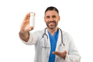 A friendly doctor with a beard and stethoscope, wearing a white lab coat, smiles while holding a small medicine bottle in a studio setting. photo