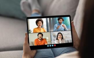 A woman sits on a couch, relaxing with her legs up, and is participating in a conference call on a tablet. The screen displays four people, with three of them in separate frames. photo