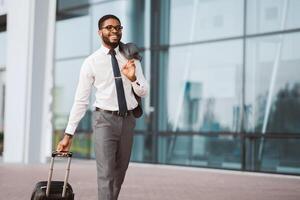 African American man in a suit is walking with his luggage at an airport. The setting is modern and busy, highlighting his professional demeanor and readiness for business travel. photo