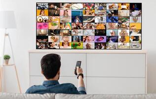 A man sits on a couch in a living room, watching television. The television screen shows multiple channels playing simultaneously, displaying a variety of content photo