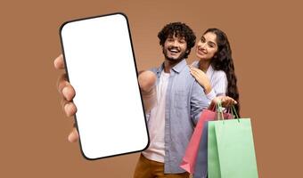 A young Indian couple holding shopping bags and a smartphone with a blank screen. The man is holding the phone out towards the camera, and the woman is standing behind him photo