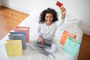 A Hispanic woman is seated on a bed, holding a credit card in one hand and a laptop in the other, possibly making an online purchase or managing finances, top view photo